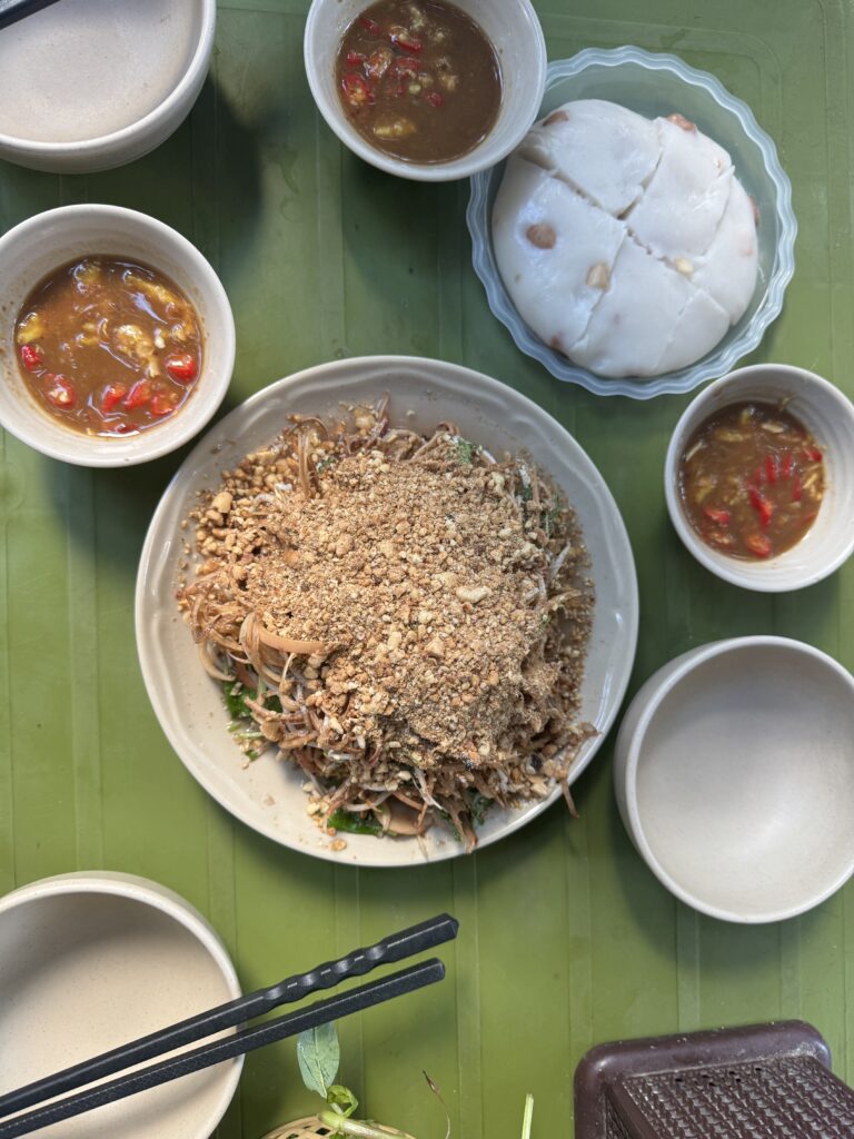 A plate of shredded chicken salad topped with crushed peanuts, bowls of dipping sauce, rice cake, and empty bowls on a green table, with chopsticks placed nearby.