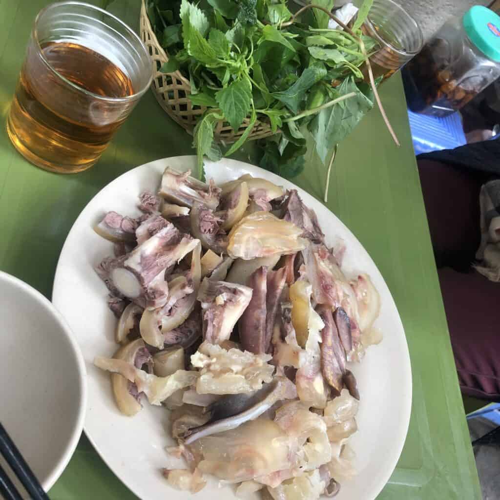A plate of sliced boiled meat and offal beside fresh herbs, two glasses of iced tea, and chopsticks on a green table.
